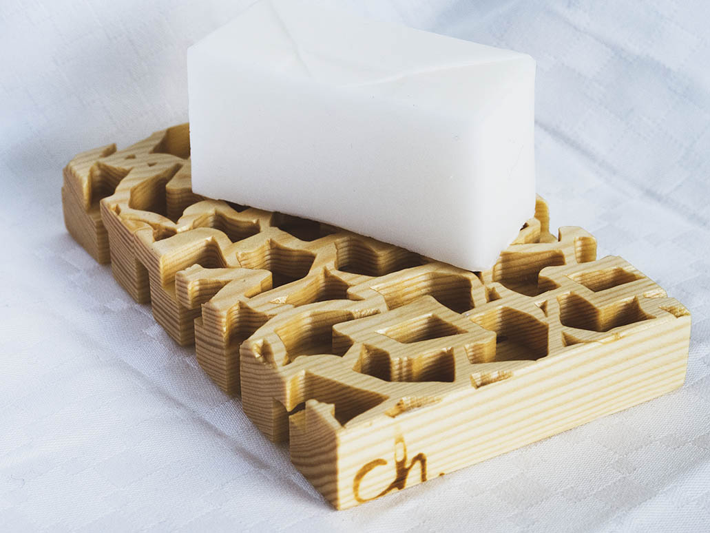 Männystä jyrsitty puinen saippua-alusta valkoista taustaa vasten. Päällä valkoinen pala saippuaa.