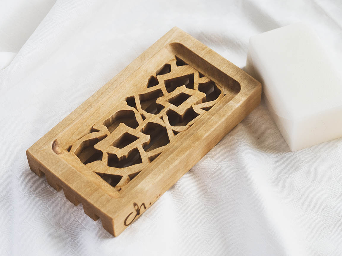 Puinen saippua-alusta valkoista taustaa vasten. Saippua-alustassa on pienet reunat ja kaksi kukkaa keskellä. Vesi pääsee valumaan rei'istä läpi pitäen saippuan kuivana.