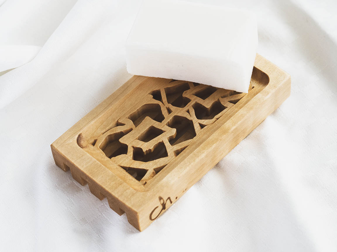 Puinen saippua-alusta valkoista taustaa vasten. Saippua-alustassa on pienet reunat ja kaksi kukkaa keskellä. Vesi pääsee valumaan rei'istä läpi pitäen saippuan kuivana.