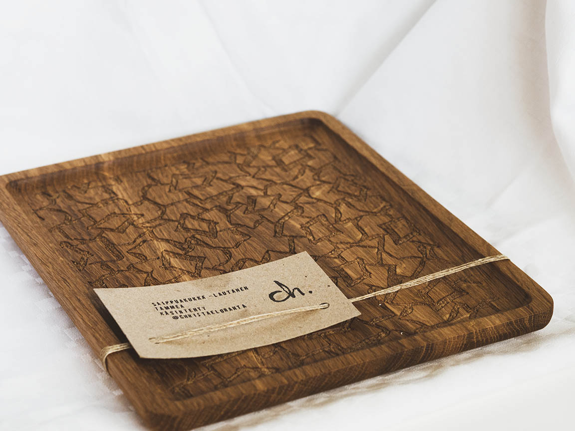 Saippuakukka-tarjoilulautanen valkoista taustaa vasten. Materiaali on tammea ja lautasta kiertää vyöte. Tarjoilulauta on neliön muotoinen, siinä on pienet reunat ja jyrsitty kukkakuvio keskellä.