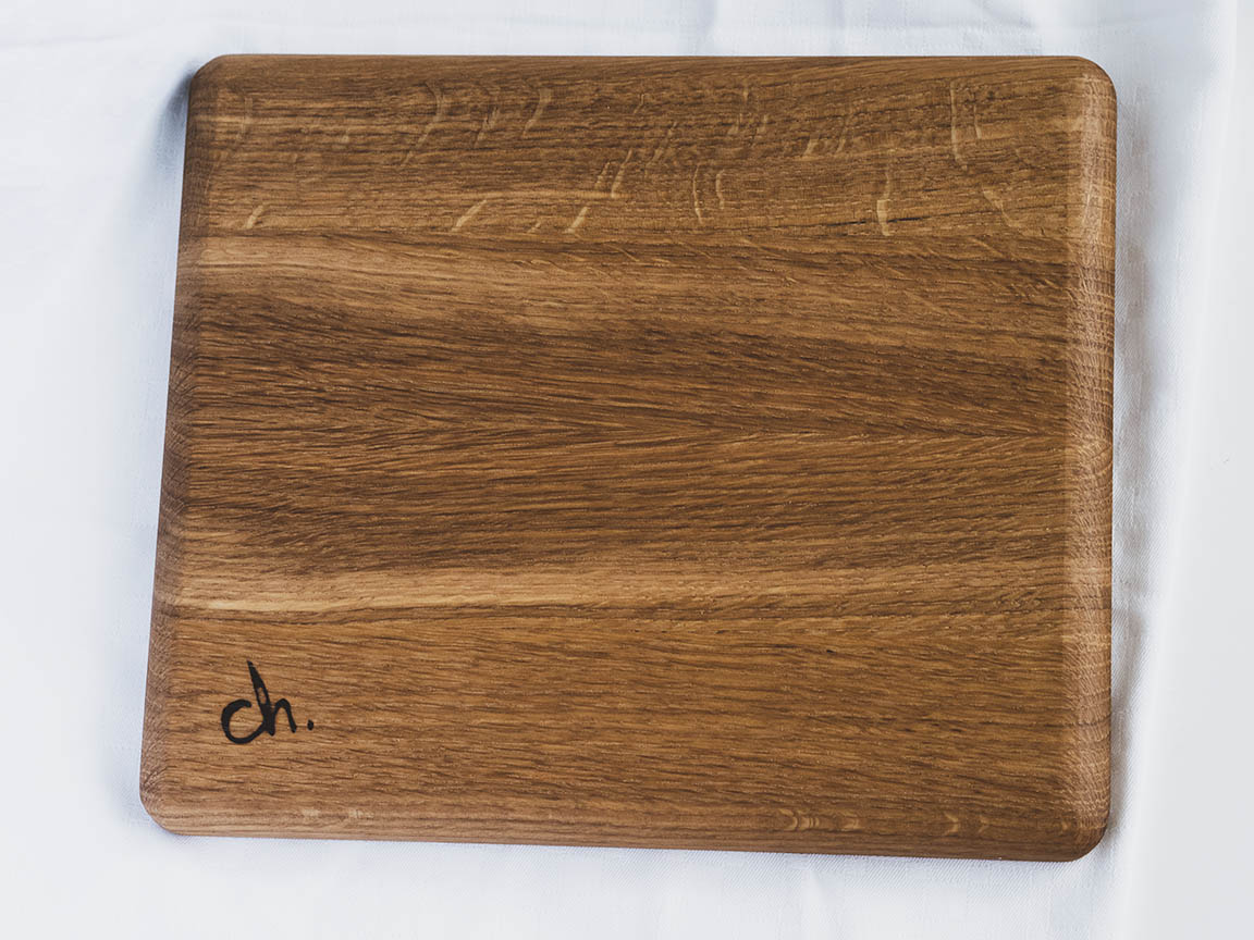 Saippuakukka-tarjoilulautanen valkoista taustaa vasten. Materiaali on tammea. Tarjoilulauta on neliön muotoinen, kuvassa lautanen on kuvattu selkäpuolelta, jossa on polttomerkittynä ch.-logo.